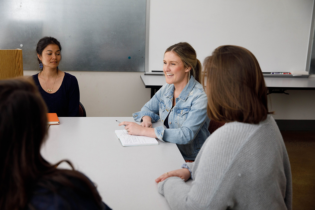 Three students listen to another student talking at a discussion table in a classroom.