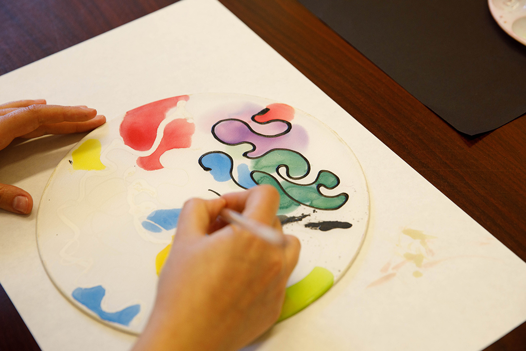 Close-up of a student's hands outlining green, purple, red, yellow and blue painted swirls on a circular surface.